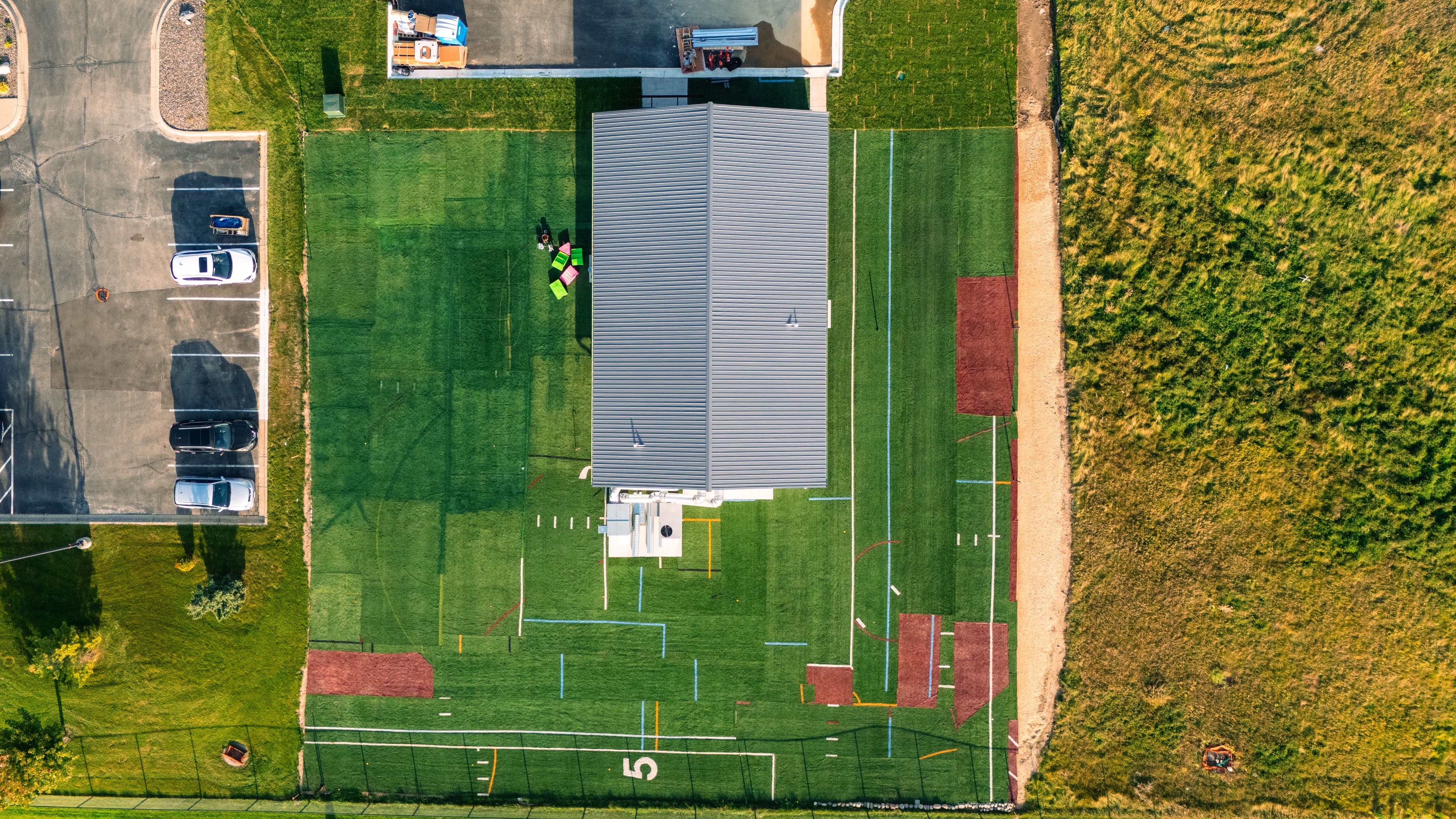 Aerial view of a sports field with a building, parking area, and green grass.