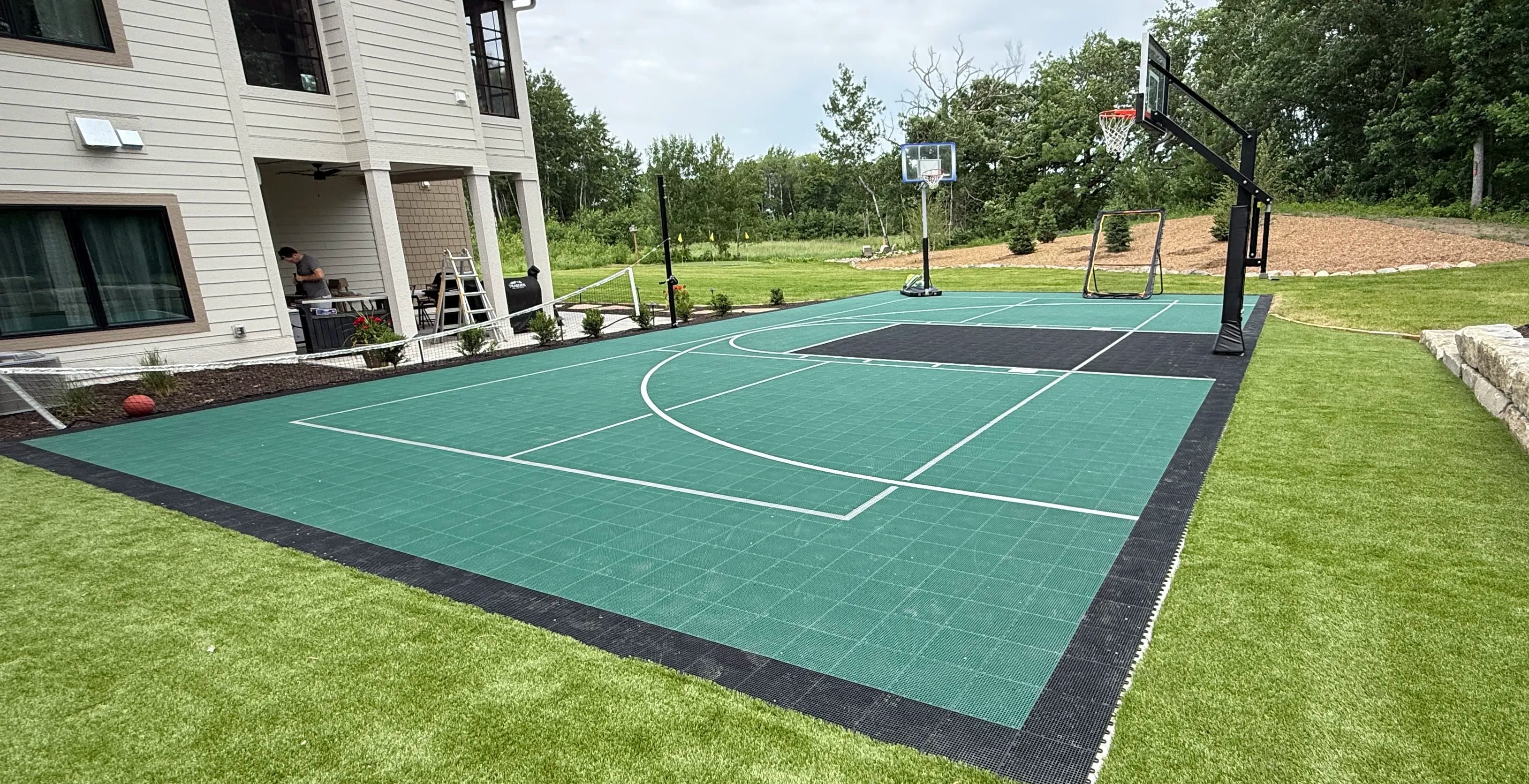 Backyard basketball court with hoop, green surface, and adjacent house, surrounded by greenery.