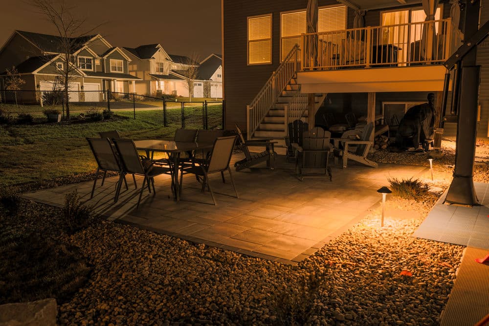 Outdoor patio at night with stone flooring, seating area, and illuminated landscaping.